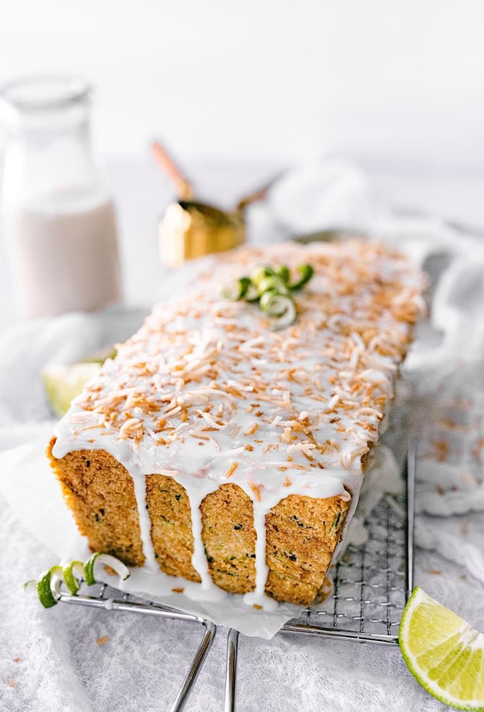 A loaf of coconut lime bread topped with white icing and toasted coconut flakes, decorated with lime zest, on a cooling rack with a jug and lime wedges in the background