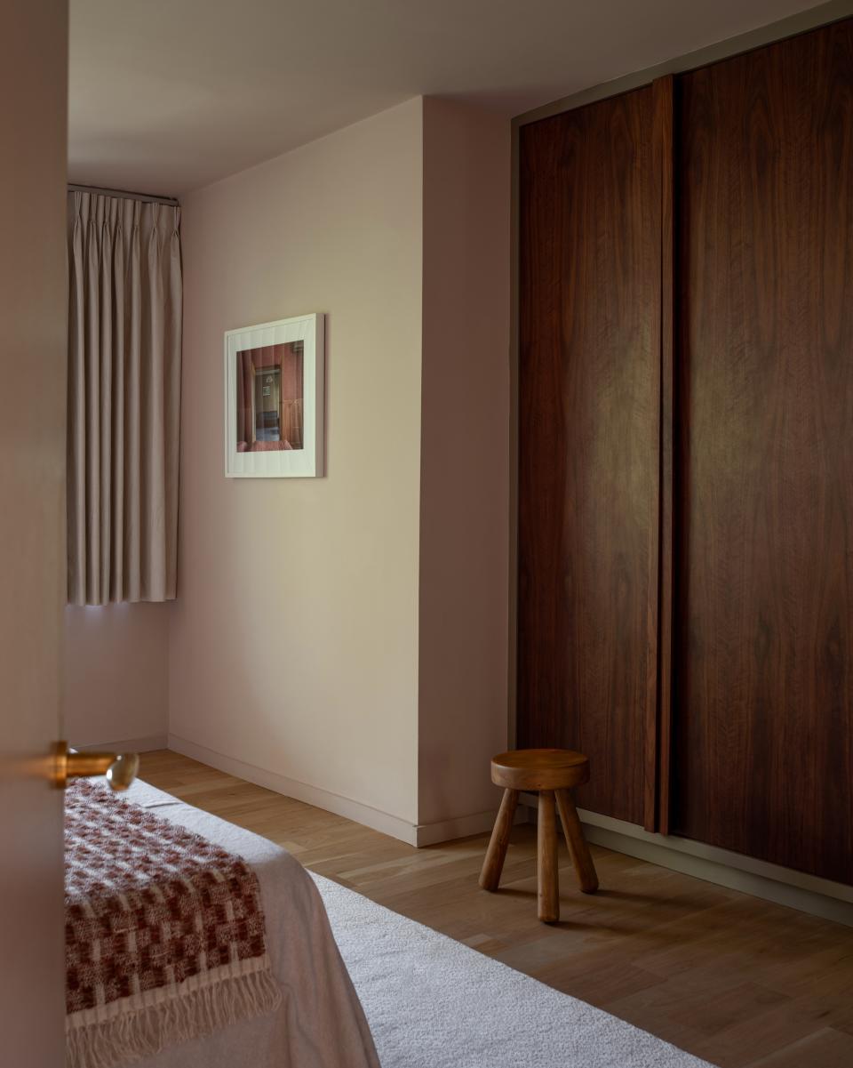 The primary bedroom is a serene oasis that basks in minimum color and maximum light. Potted Shrimp by Farrow & Ball calms the walls, while a stool by Ingvar Hildingsson echoes the walnut wardrobes. A photograph by Stephen Shore of a hotel in Ukraine emblazons the wall.