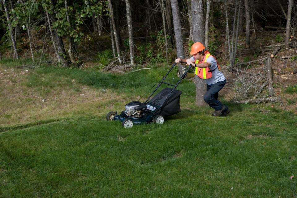 A worker in a safety vest pushes a lawn mower across a green lawn. 