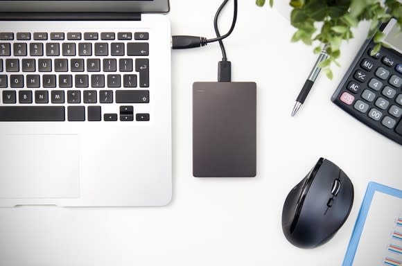 An external disk drive connected to a laptop computer on a desk with a pen, mouse, and calculator nearby.