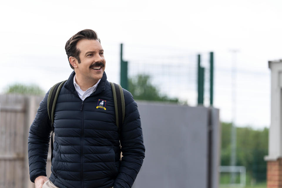 A mustachioed man in a navy puffer coat cheerfully walks through dreary london wearing a backpack (Colin Hutton / Apple TV)