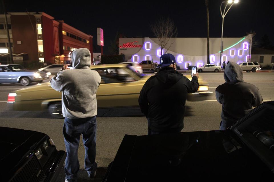 Spectators use their phones to photograph passing lowriders on a city street.