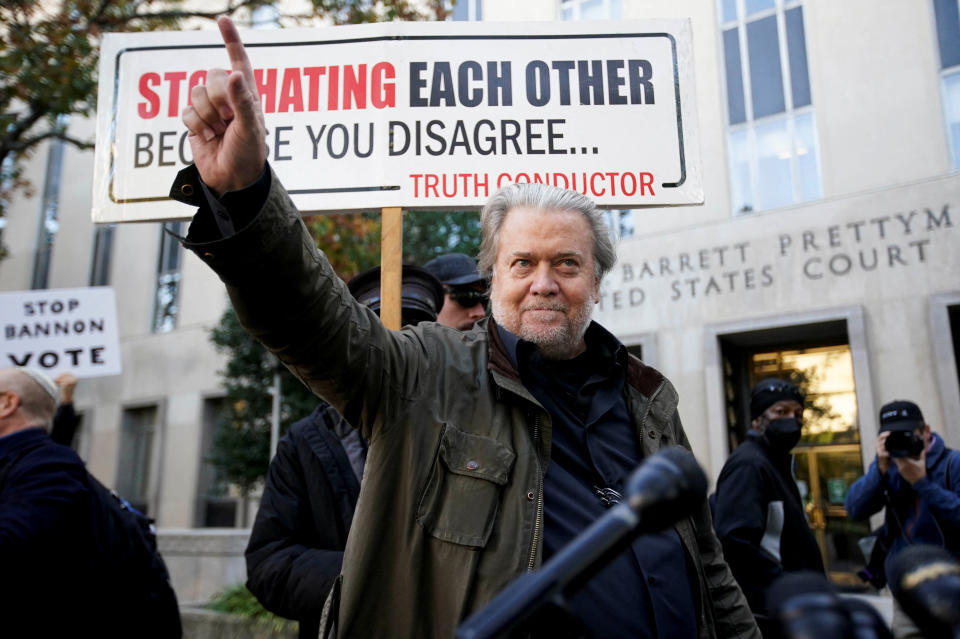 Steve Bannon raises his arm to point in front of someone holding a sign saying: Stop Hating Each Other Because You Disagree.