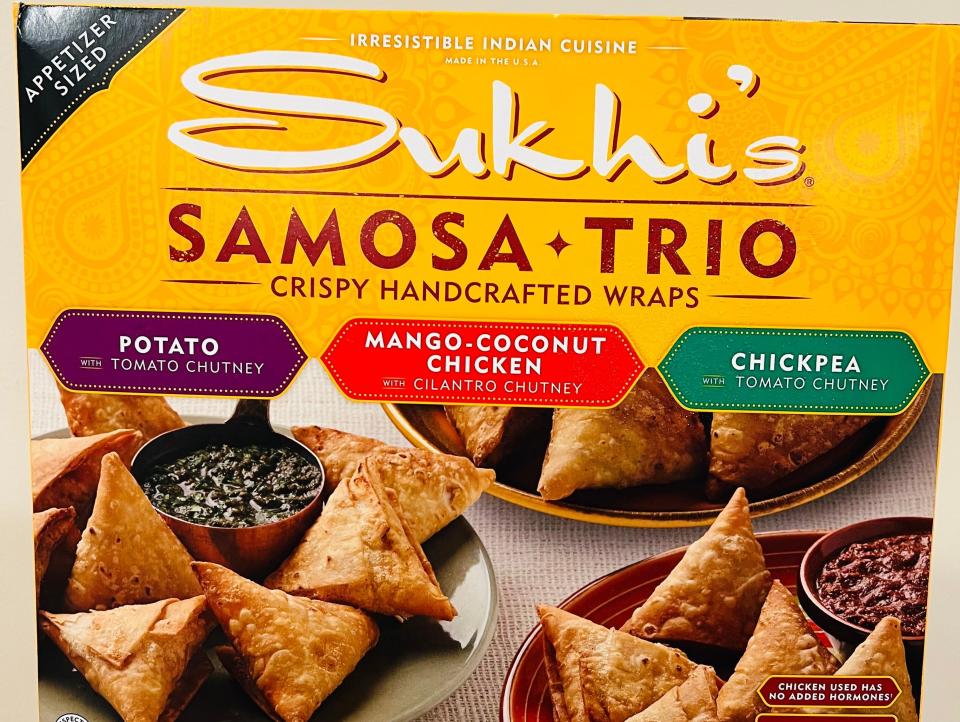 Box with yellow packaging that says "Sukhi's Samosa Trio" and three labels with "Potato," "Mango Coconut Chicken," and "Chickpea" above pictures of samosas on trays