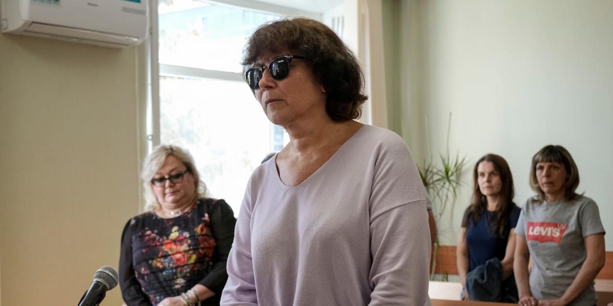 Irina Tsybaneva, 60, stands wearing sunglasses and a lavender sweater in a courtroom in St. Petersburg, Russia, Thursday, May 11, 2023, convicted of desecrating the grave of Putin's parents. Three other people stand in the background.