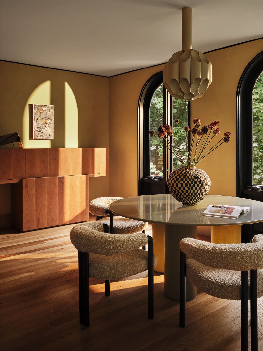 in a bar and lounge area is a wood floor, arched windows, a walnut sideboard, a round table with three boucle covered low back chairs, a multifaceted round pendant, and a checkerboard patterned vase