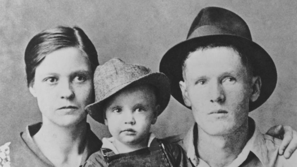 a black and white photo of a man and woman posing with a child, who wears overalls and a fedora