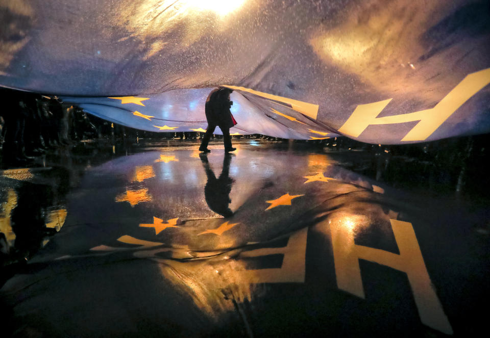 FILE - In this Saturday, Jan. 20, 2018 file photo a protester walks under a large European Union flag bearing the words "Here_Now" during a protest outside the parliament building in Bucharest, Romania. Tens of thousands of Romanians on Saturday protested against legislation passed by Parliament which critics say will make it harder to prosecute crime and high-level corruption, briefly scuffling with riot police as they massed in University Square. (AP Photo/Vadim Ghirda, File)