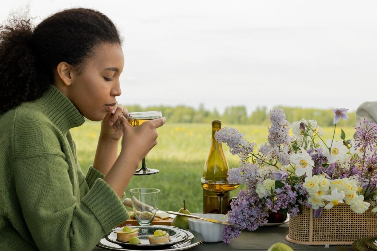 Black woman in green turtle neck drinking wine on a vinyard while eating grapes and cheese