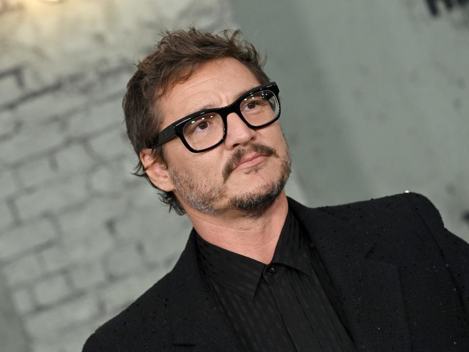 pedro pascal frowns in thick dark rimmed glasses and dark suit