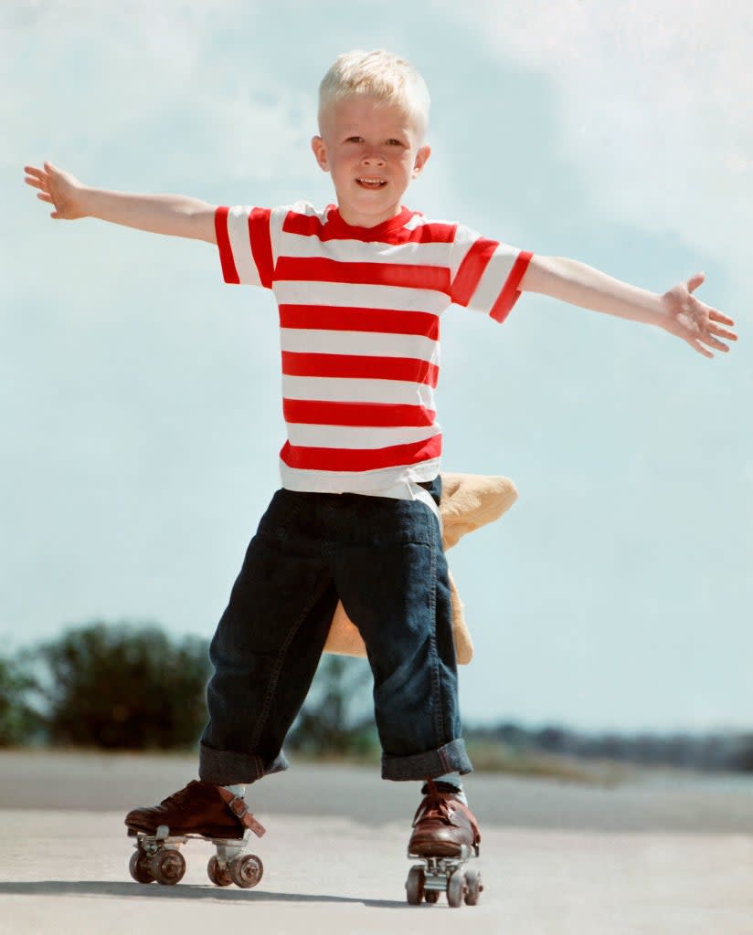 A young boy smiles and balances on roller skates with arms outstretched. He is wearing a striped shirt and denim pants. The background appears to be outdoors