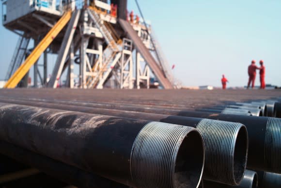 A close-up of drill pipes with oil workers and a rig in the background.