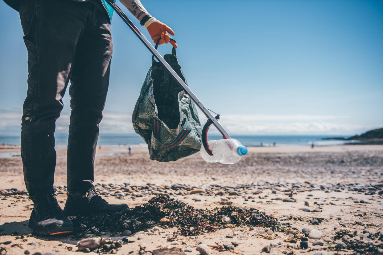 A person holding a litter picker with a plastic bottle in it and a bag on a beach