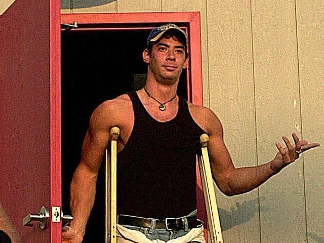 Eddie walking out a door in a baseball hat, tank top, and crutches.