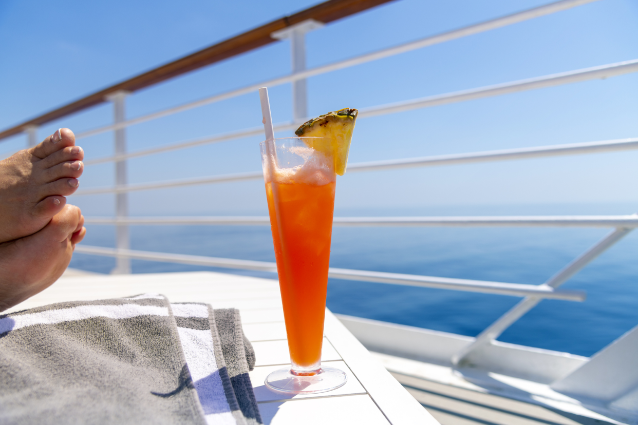 Focus on orange tropical cocktail drink with pineapple on cruise ship, on a white table with feet to the left side, blurred background of deck and ocean