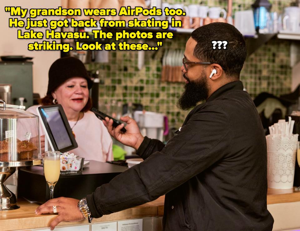 A man wearing a black jacket stands at a coffee shop counter, talking to a woman behind the counter. The counter has pastries and drinks displayed