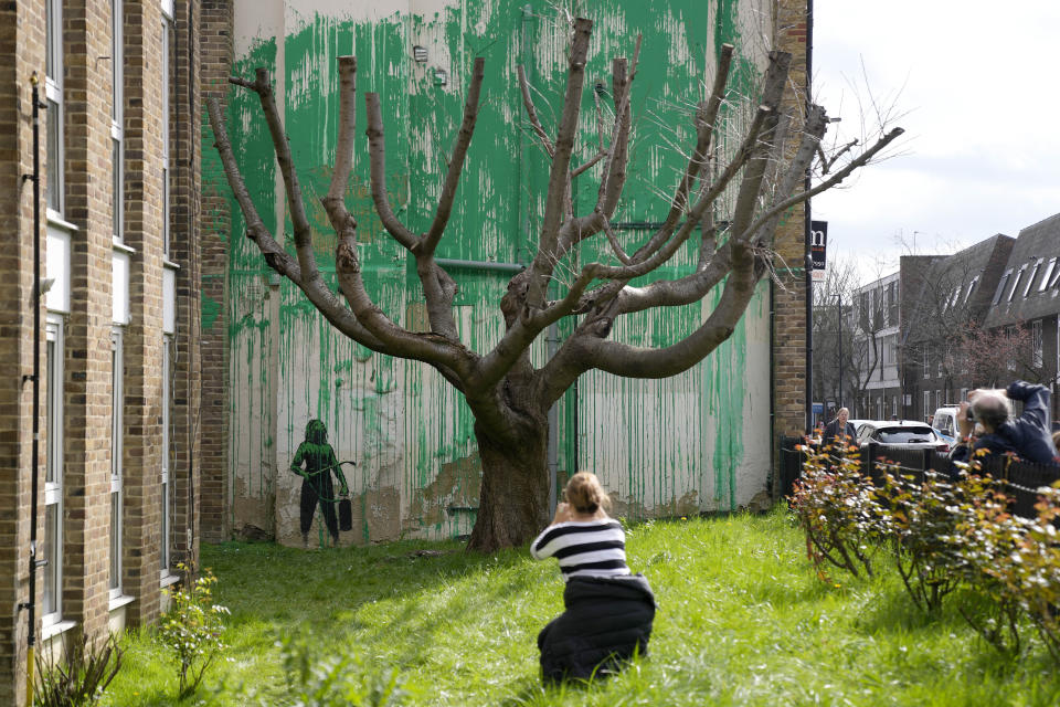 People look at a new Banksy painting on a wall in London, Monday, March 18, 2024. A new Banksy mural drew crowds to a London street on Monday, even before the elusive graffiti artist confirmed that the work was his. The artwork in the Finsbury Park neighborhood covers the wall of a four-story building and shows a small figure holding a pressure hose beside a real tree. Green paint has been sprayed across the wall, replicating the absent leaves of the tree, which has been severely cropped. Banksy claimed the work by posting before and after photos of the location on his official Instagram account.(AP Photo/Alastair Grant)