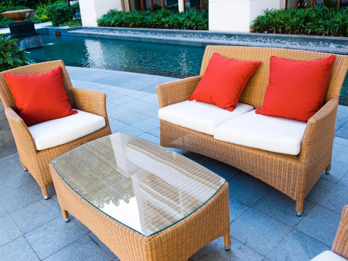 light brown outdoor wicker furniture set with red pillows and a pool in the background