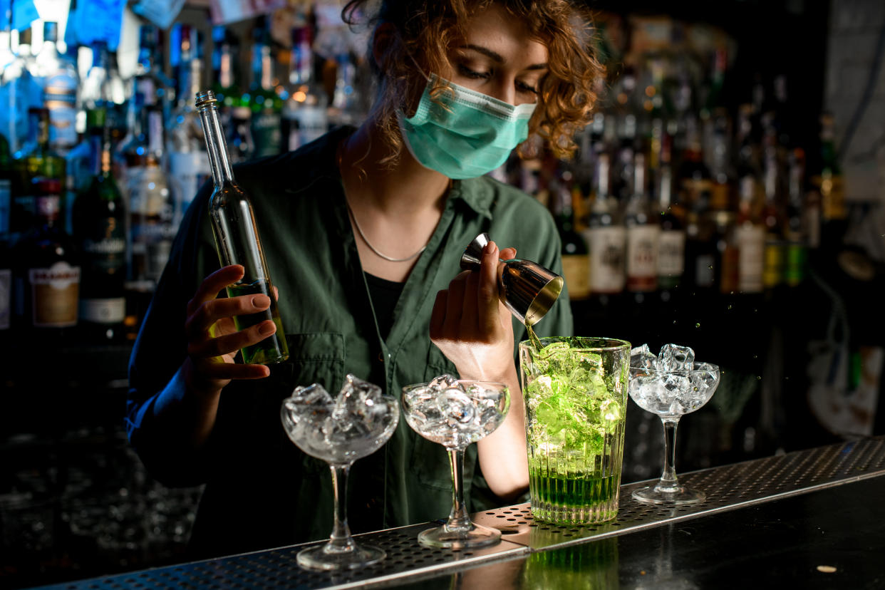 Young bartender girl in a medical mask pour green liquid from beaker into glass with ice. Medical mask for prophylaxis and protection from coronavirus COVID-19.