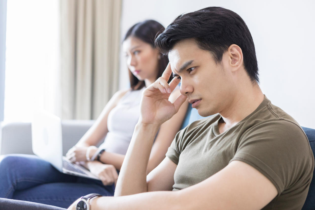 A mid-adult man puts his hand up to his head and brainstorms, while his wife sits in the background with a laptop.