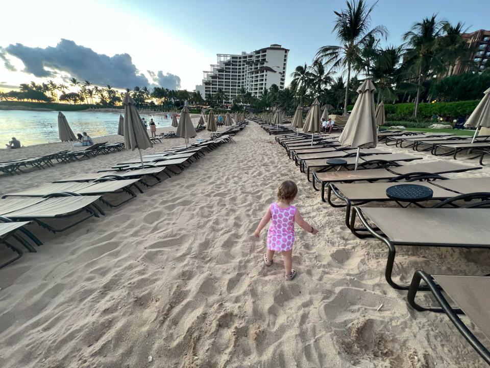 A child in a white and pink dress walking on the sand of a beach in between rows of beach chairs.