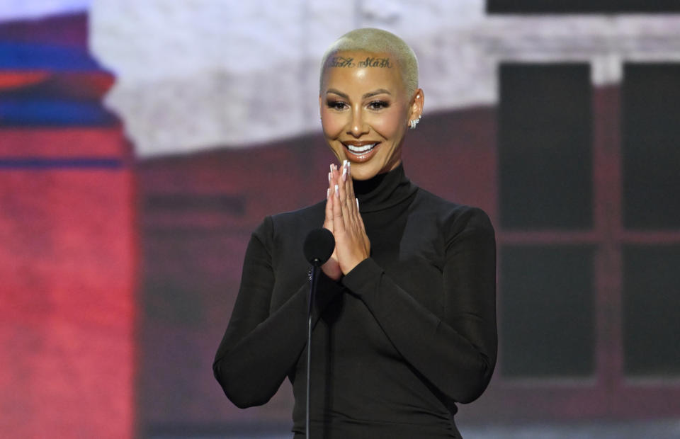 Amber Rose smiling while speaking at podium, hands clasped together. She wears a sleek turtleneck dress and has shaved hair