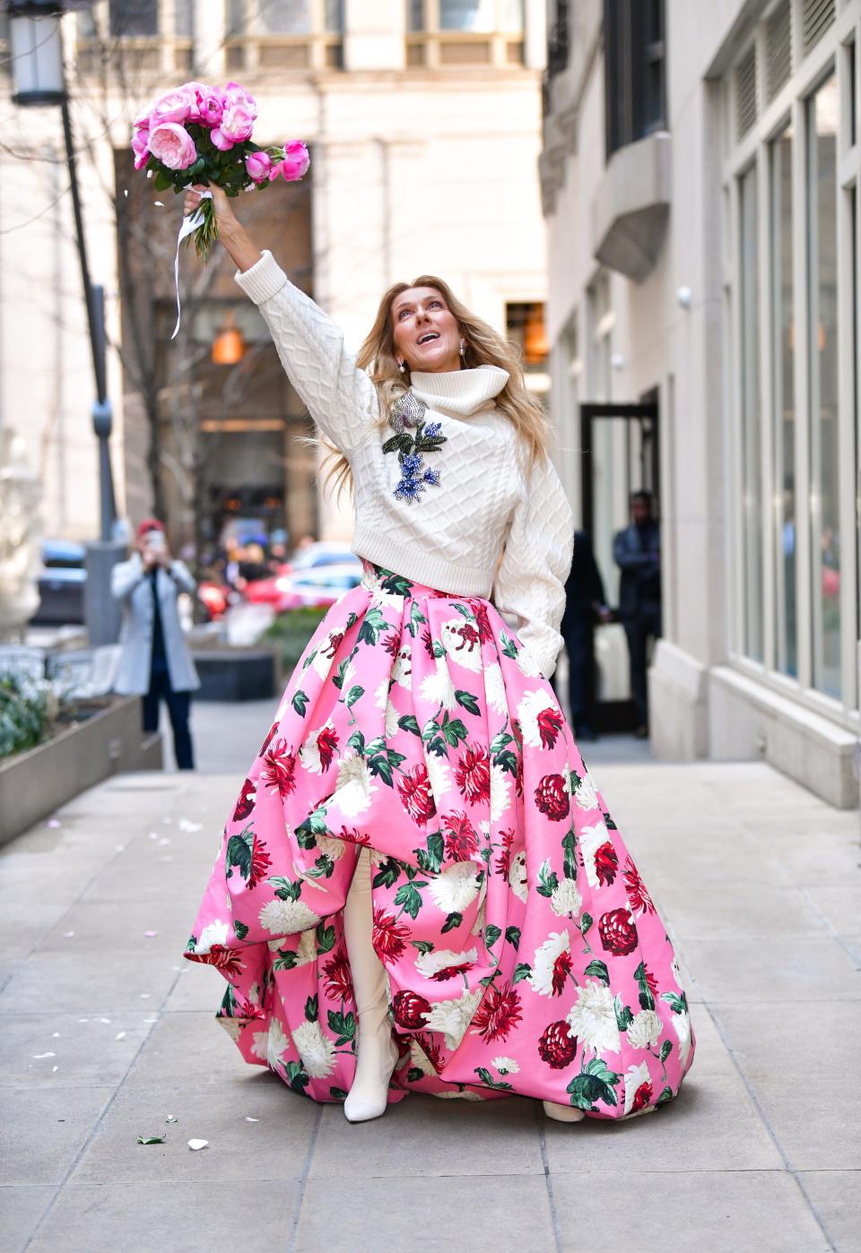 Celine in a cream sweater with a blue flower on it, a voluminous pink and floral print skirt, and tall white boots. She's holding up a matching pink bouquet of flowers.