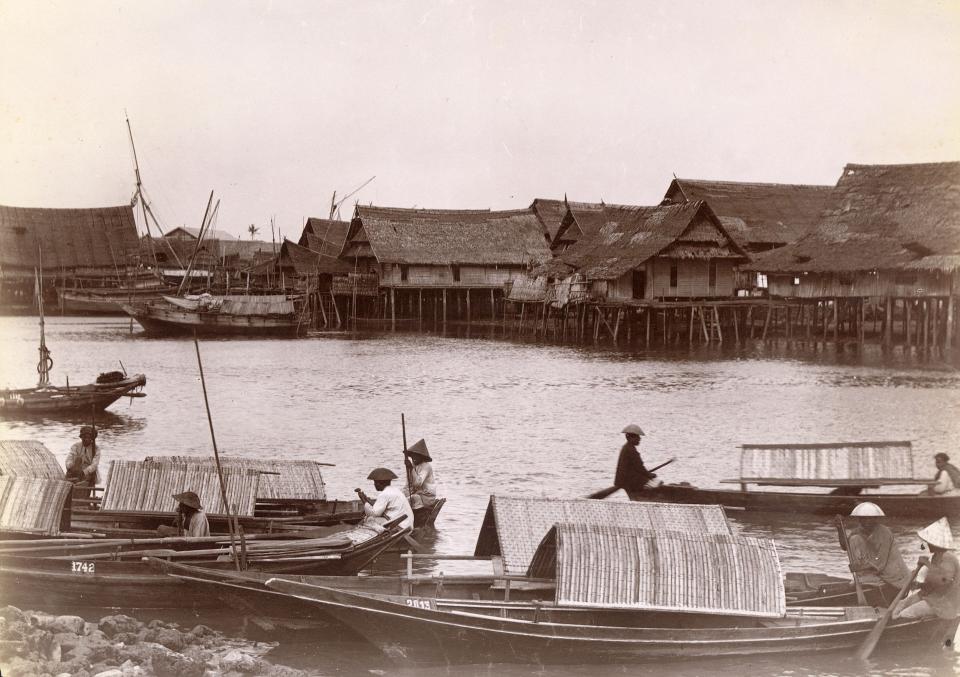 A vintage photo of sampan boats floating near houses on stilts on the water.