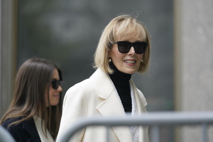 E. Jean Carroll, in cream coat and dark glasses, smiles over at a photographer behind a barrier.