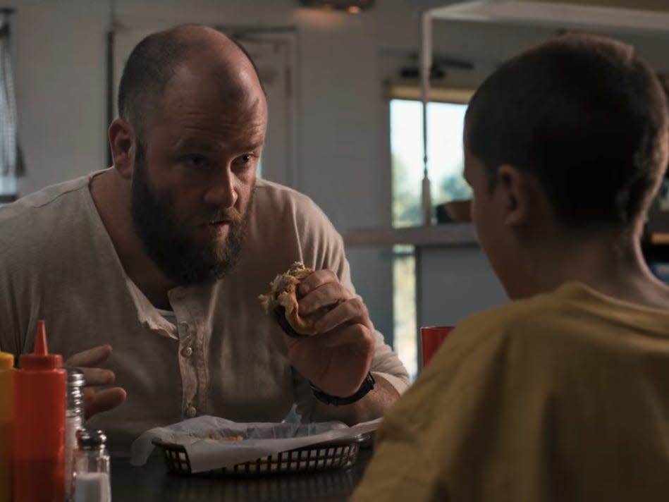 a man with a bald head and beard holding a hamburger in stranger things, sitting across from a young girl with a buzz cut