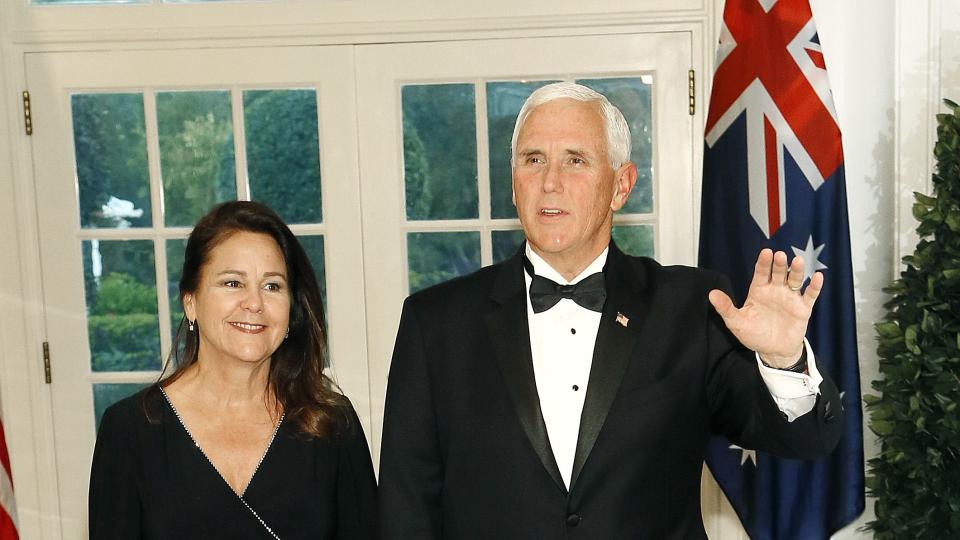 karen and mike pence walk in a hallway, they both wear black formal attire and hold hands as mike pence waves with his free hand