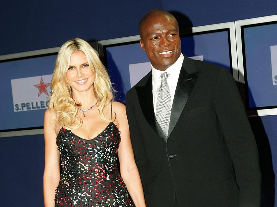 Heidi smiling in a black green and red dress and Seal smiling in a black suit.
