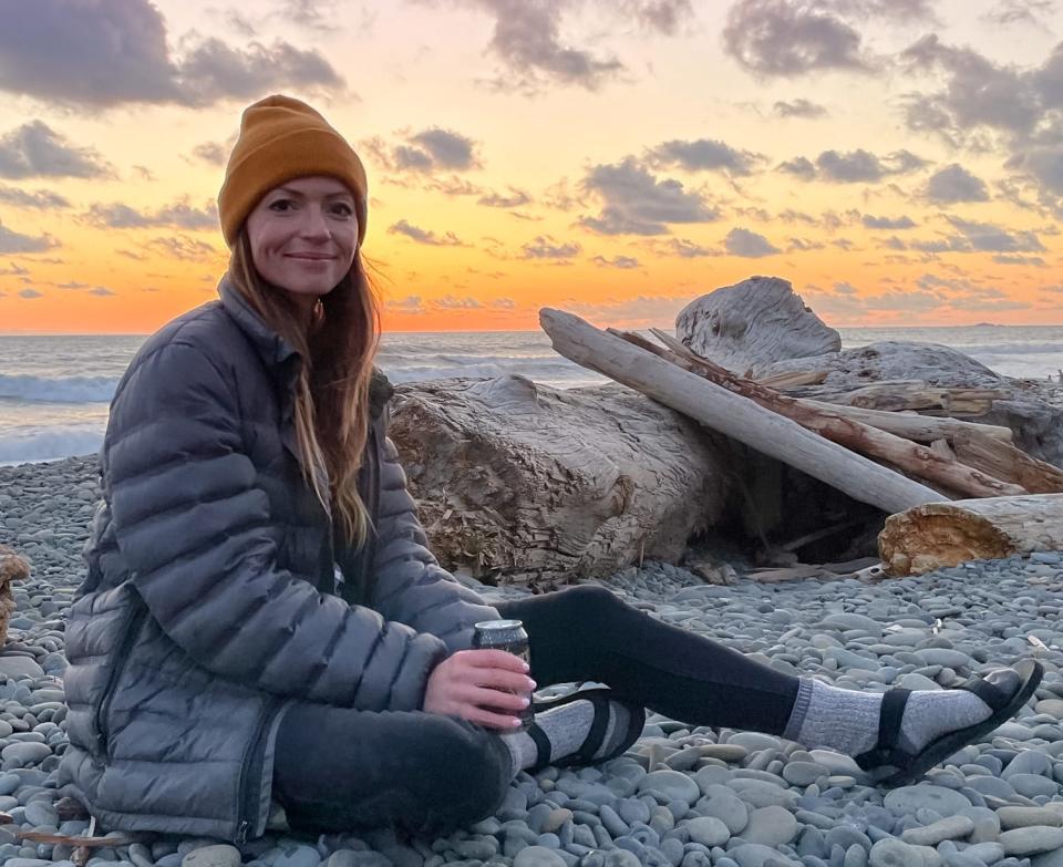 Emily, wearing a beanie, puffer jacket, black leggings, high socks, and sandals, sits on a rocky beach near driftwood at sunset.