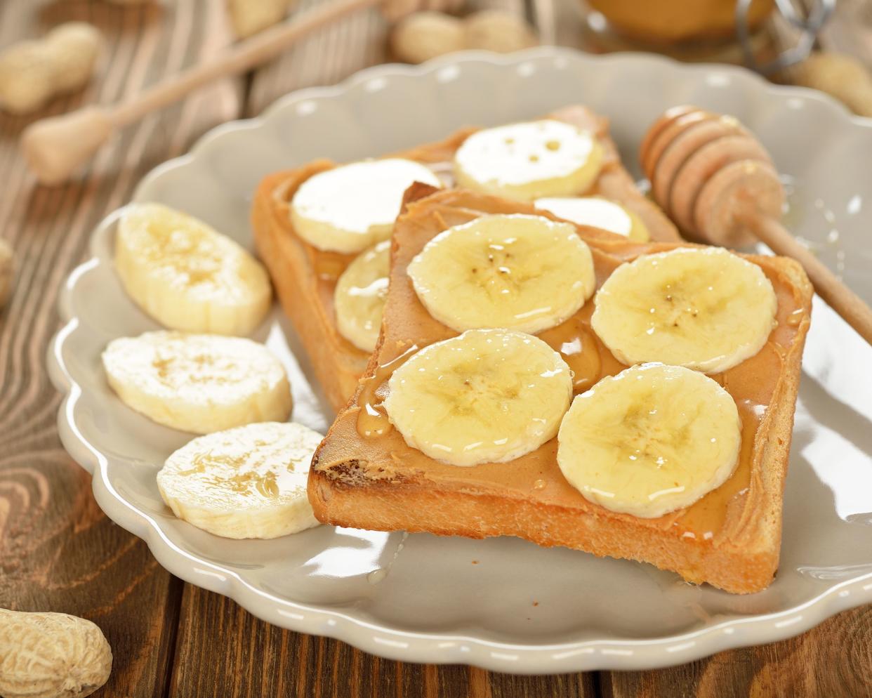 Two slices of peanut butter and banana toast with a honey dipper on a white plate on a wooden table with a blurred background of ingredients