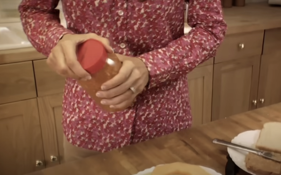 A person in a patterned shirt is opening a jar of peanut butter in a kitchen. A sandwich and a knife are visible on the counter