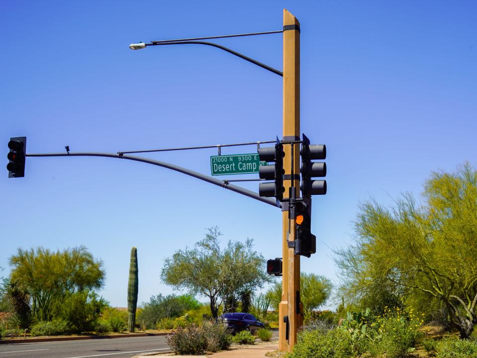 A street sign at a stoplight says Desert Camp drive with cacti, trees, and blue skies in the background