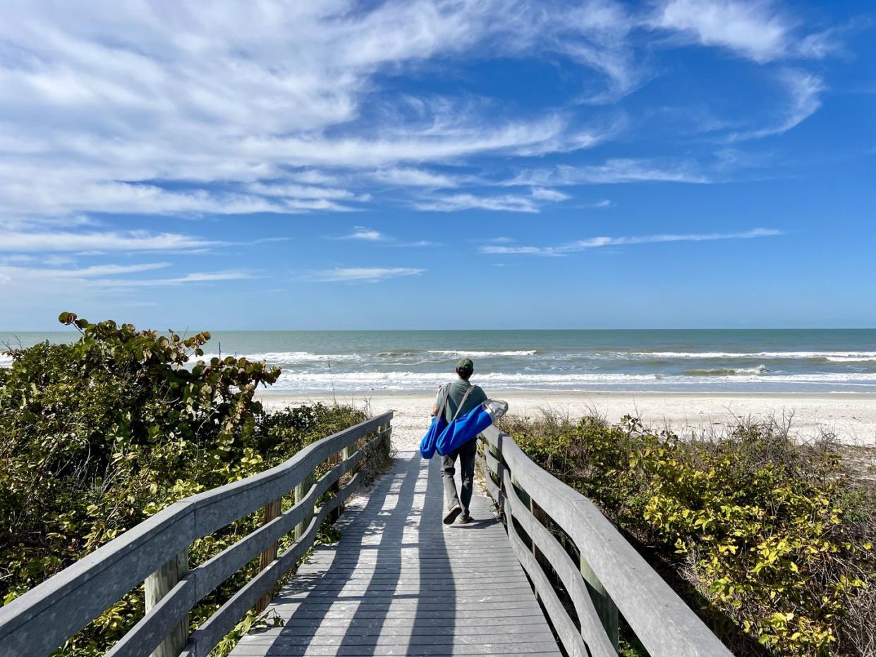 senior man carrying beach chairs over footpath against sand, water and blue skies