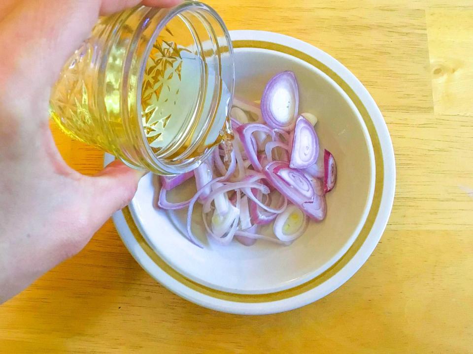 A hand holding a jar of oil pouring into a small white bowl of sliced shallots on a wooden table