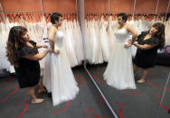 In this Friday, June 21, 2019, photo Ann Campeau, left, owner of Strut Bridal, fits a new dress on inventory manager Stefanie Zuniga at her shop in Tempe, Ariz. Cut-rate prices on websites that sell wedding dresses direct from China put pressure on Campeau, who owns four bridal shops in California and Arizona. She has had customers come in after seeing low-end gowns online and expecting to get a dress at a similar price. (AP Photo/Matt York)