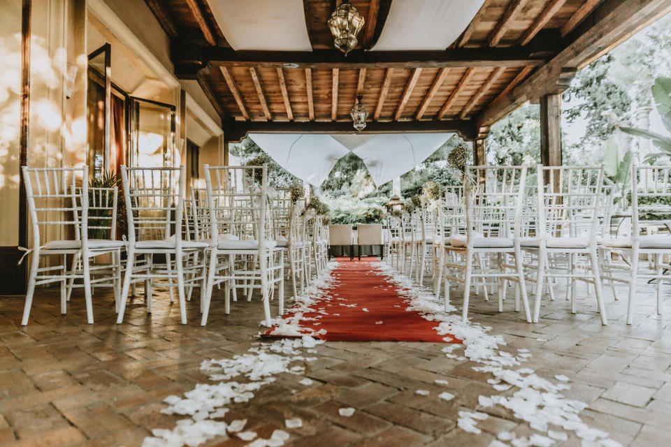 Wedding ceremony setup with white chairs arranged in rows along a red aisle runner scattered with white petals, under a covered outdoor structure