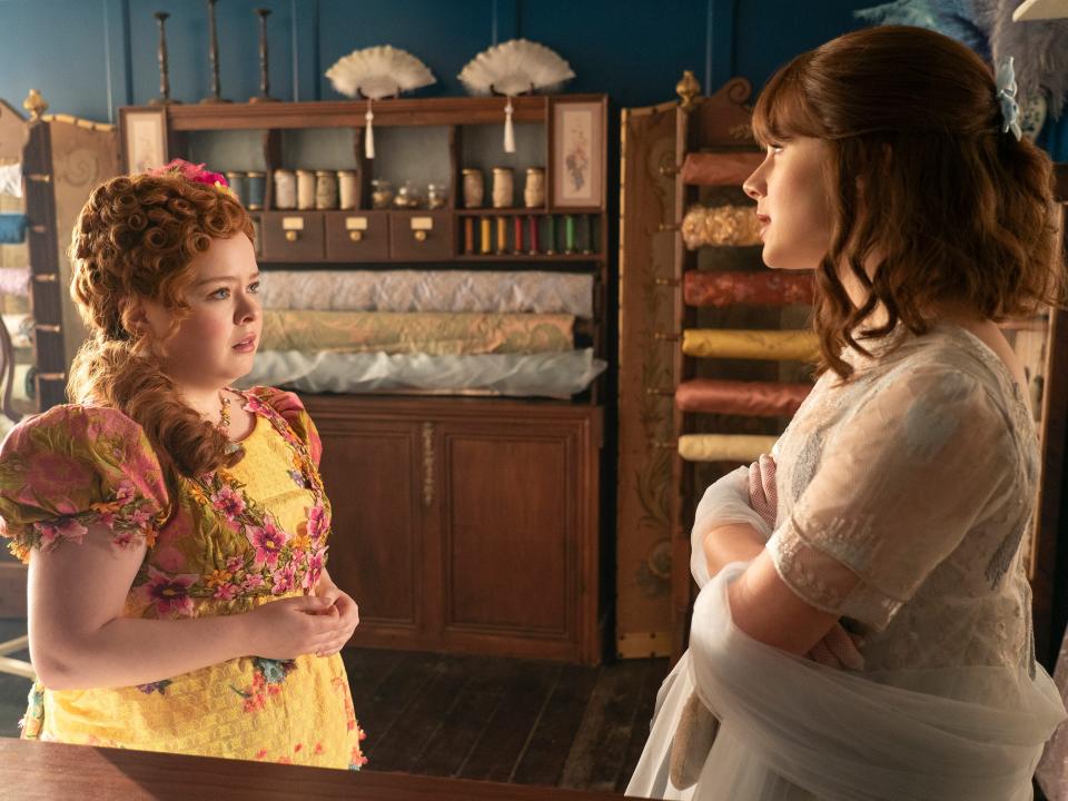 penelope featherington, in a bright yellow dress, facing eloise bridgerton, in a sheer blue gown, standing inside the modiste's stop