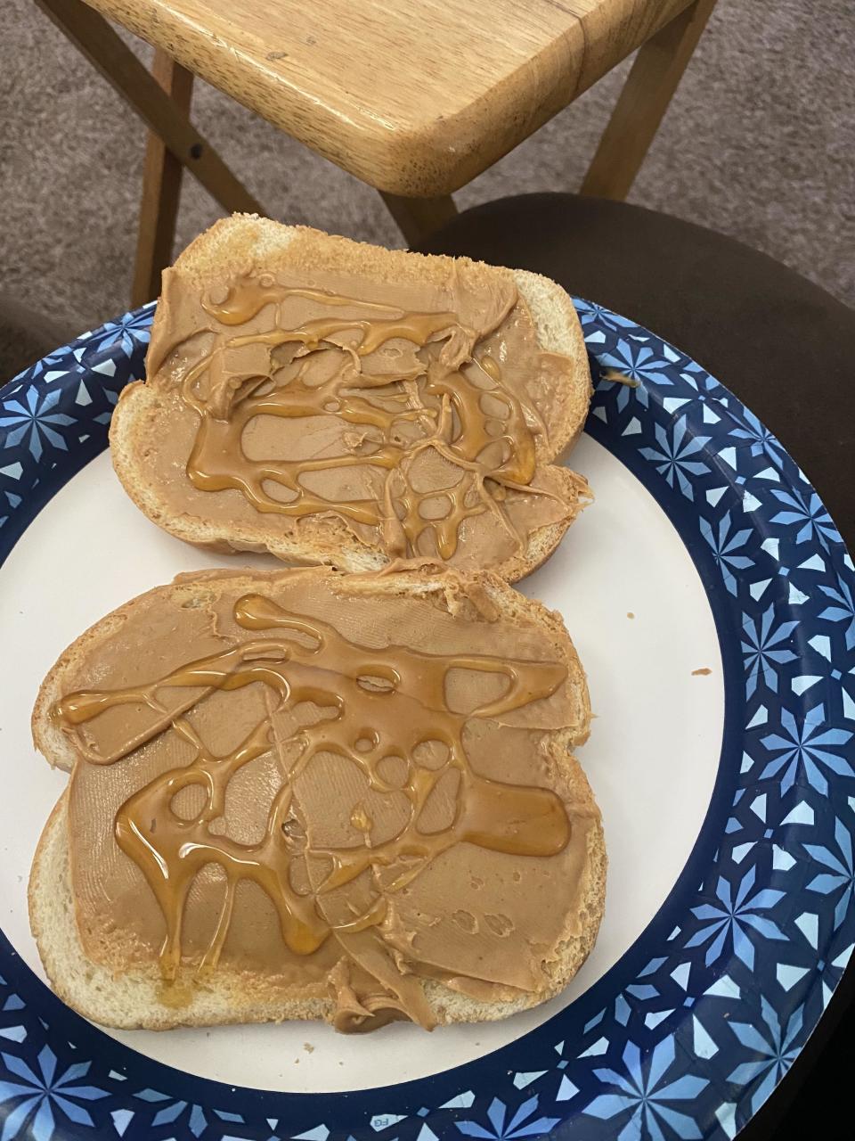 Two slices of bread with peanut butter and honey on a blue and white patterned paper plate, placed on a wooden tray table