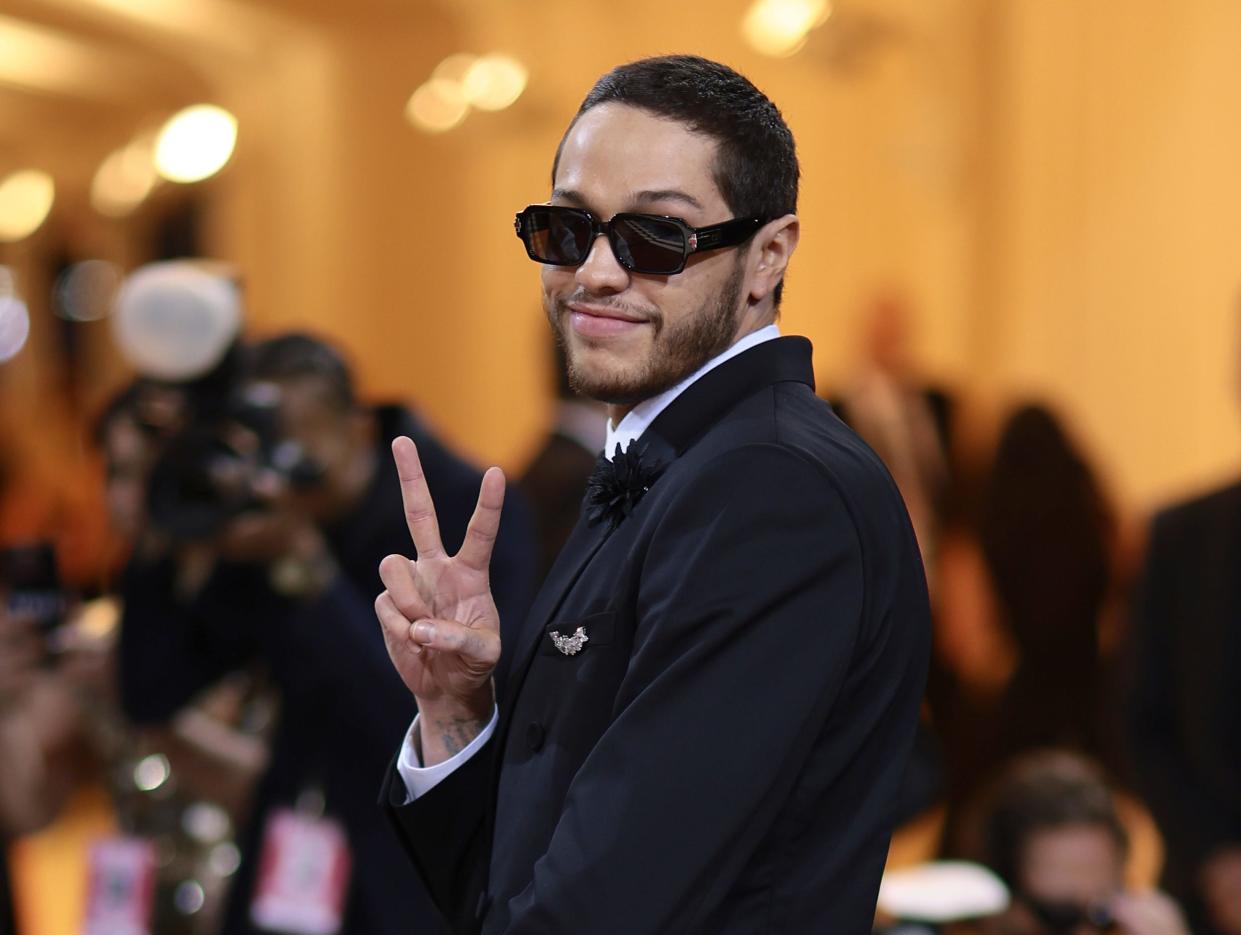 Pete Davidson holding up a peace sign with his right hand while wearing blacked-out sunglasses and a black suit in a brightly light room.