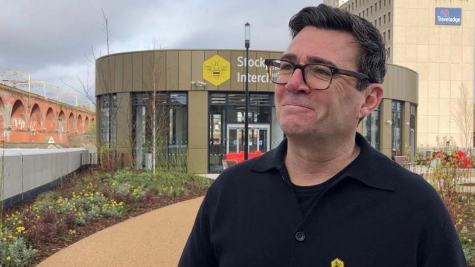 Andy Burnham smiles at the camera outside Stockport Interchange. He has dark short hair and is wearing dark rimmed glasses and a dark shirt and t-shirt. Railway arches and a Travelodge are behind him.