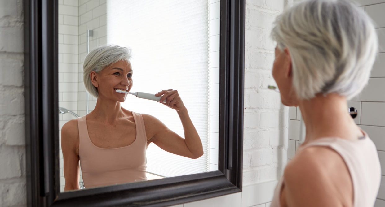 woman with short grey hair brushing teeth in mirror wearing tank top