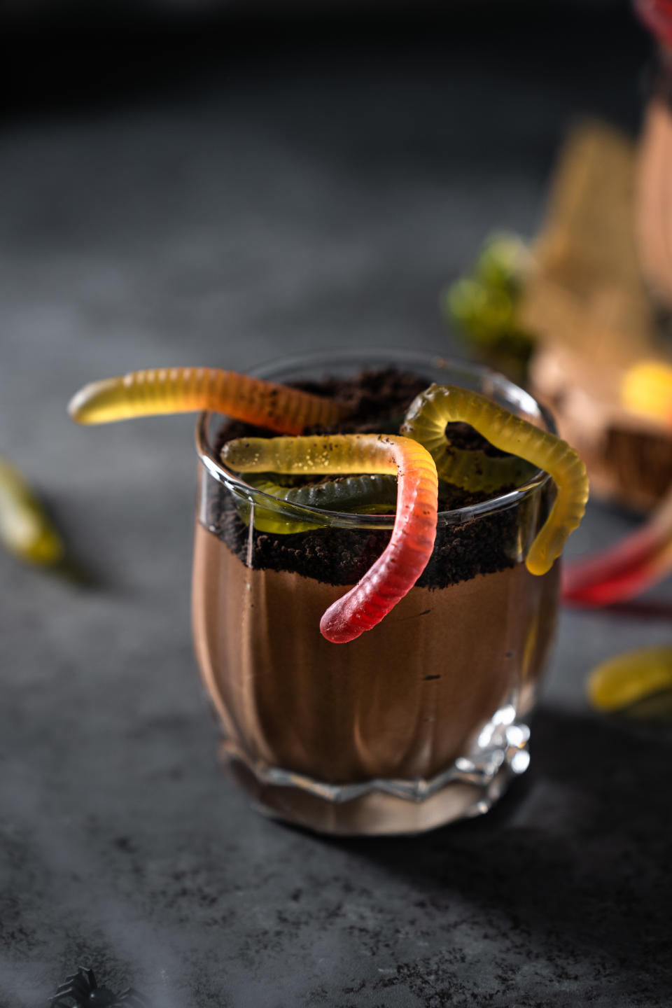 A clear glass contains chocolate mousse topped with crushed Oreos and gummy worms, resembling dirt with worms. A fun, Halloween-themed dessert