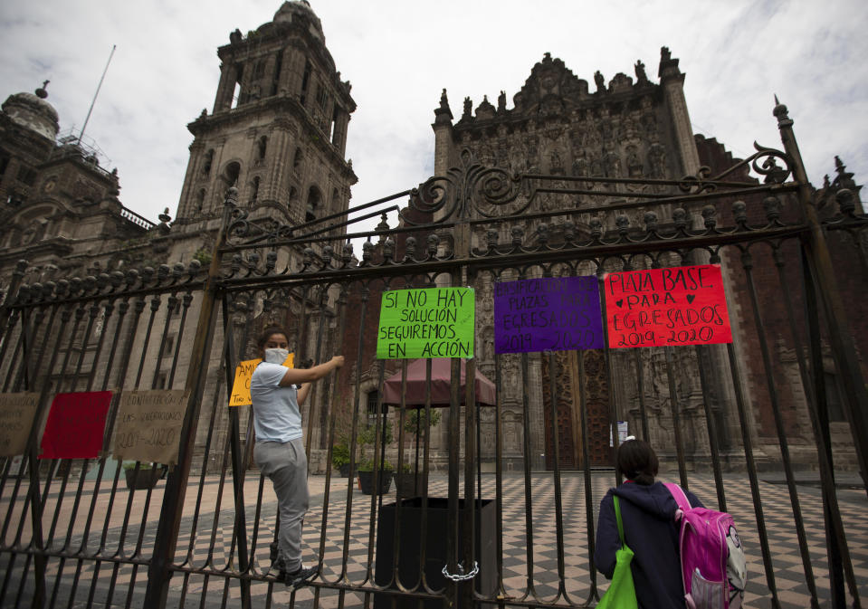 Newly graduated teachers from the state of Michoacan, hang signs on the fence of the city's cathedral, as they protest against Mexico's President Andres Manuel Lopez Obrador, demanding better salaries and jobs, in Mexico City, Thursday, Aug. 13, 2020. With the country facing a deep economic recession, Mexican President Andres Manuel Lopez Obrador has pushed to reopen the economy quickly even as COVID-19 infections and deaths continue to rise. (AP Photo/Fernando Llano)