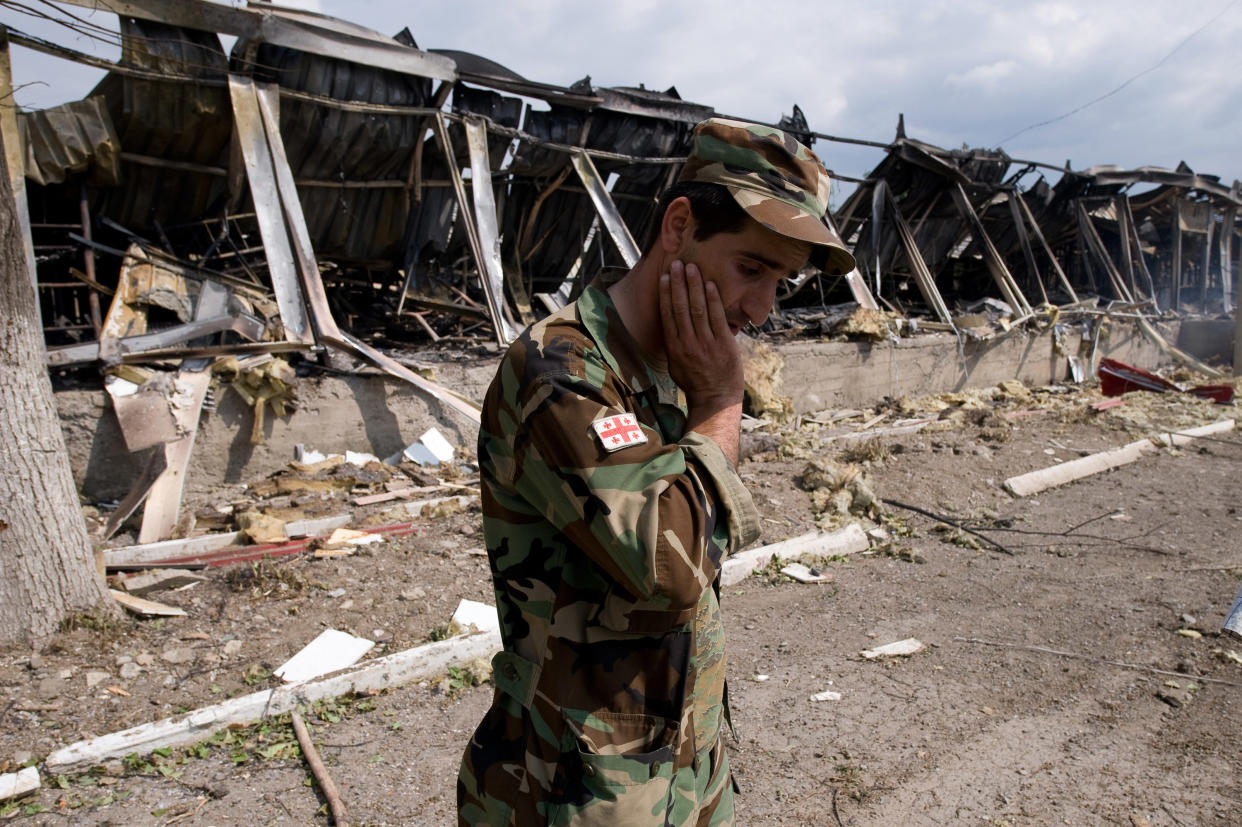 A Georgian soldier with his hand on his face looks down at the ground amid debris near a damaged building.