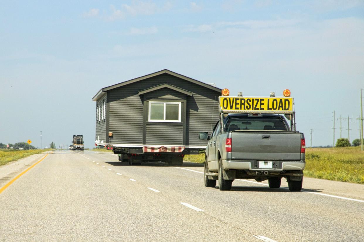 flatbed truck transporting a tiny home on trailer on a highway during a clear summer day, a truck driving behind it with 'Oversize Load' sign