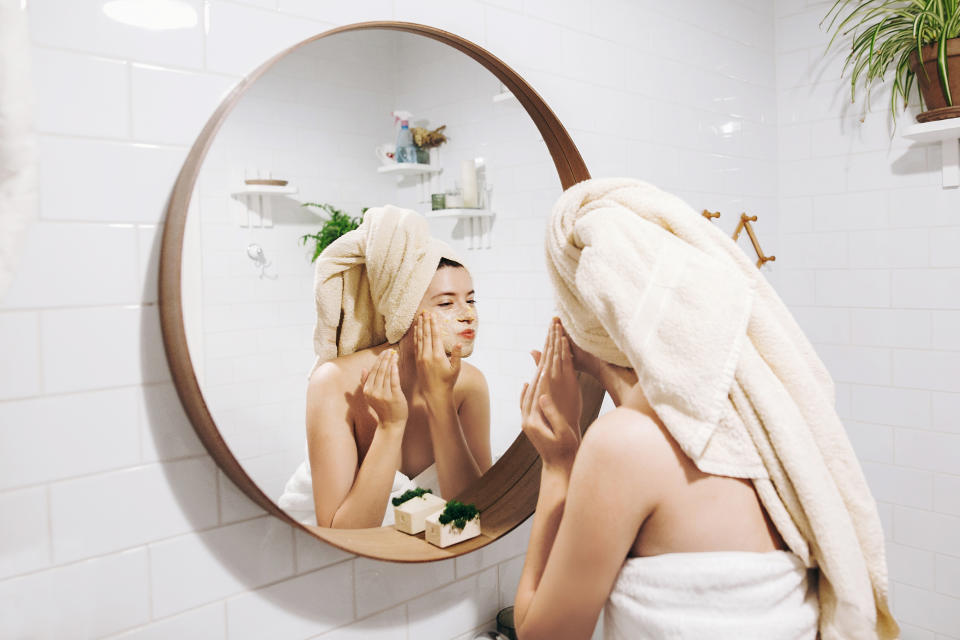 Young happy woman in towel making facial massage with  organic face scrub and looking at mirror in stylish bathroom. Girl applying scrub cream, peeling and cleaning skin. Skin Care